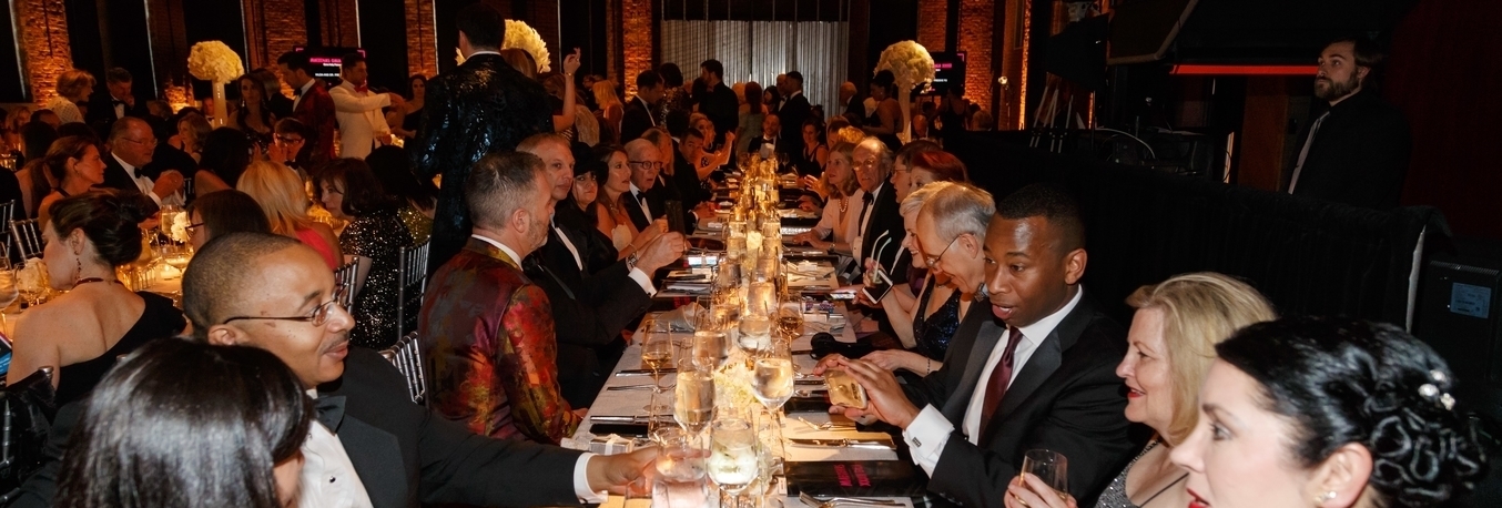 A long table with men and women in dresses and suits sitting on both sides and converse with each other