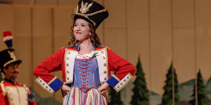 A woman in a colorful dress wears a guards jacket and hat with her hands on her hips like Wonder Woman and smiles with her mouth closed