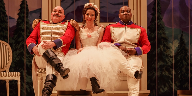A woman in a wedding dress and black boots sits on a couch with two bald guards sitting next to her. They are all smiling 