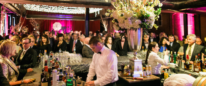 A large crowd of well-dressed men and women stand around a bar. A male and female bartender make drinks. In the middle of the bar is a large silver vase with large bouquet of white flowers