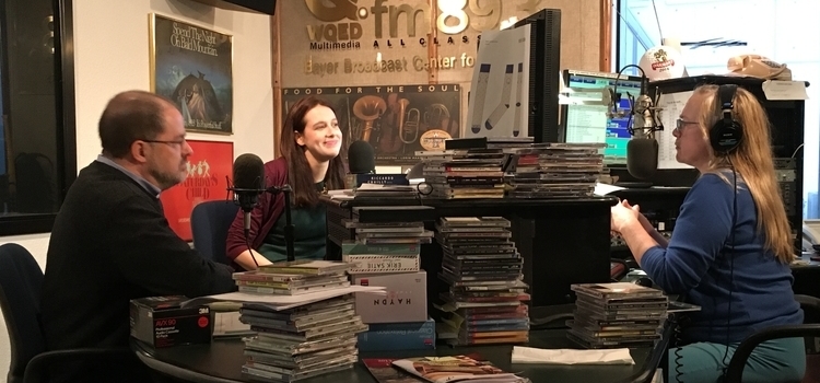 A woman sits at a messy desk with stacks of CDs and speaks into a microphone. A man and a woman sit across from her and speak into a microphone
