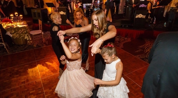 A woman in a dress spins two little girls in dresses while people in the background dance