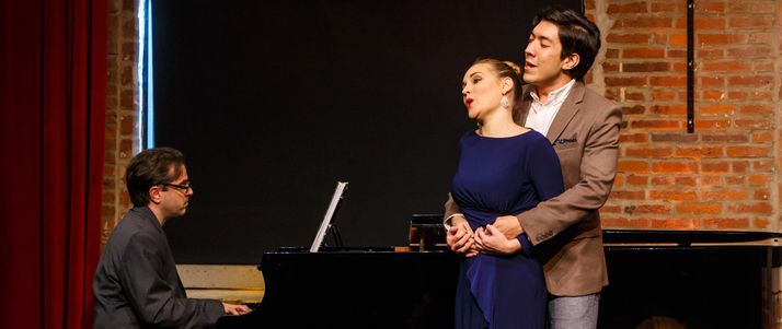 A man plays the piano while a man and woman sing.The singing man hugs the woman from behind. 